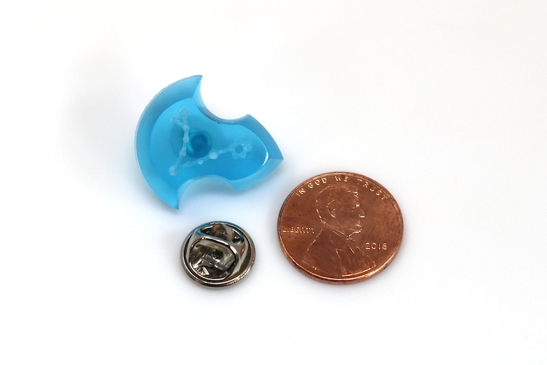May include: A blue resin pin with a white outline of the constellation Pisces. The pin is about the size of a US penny.