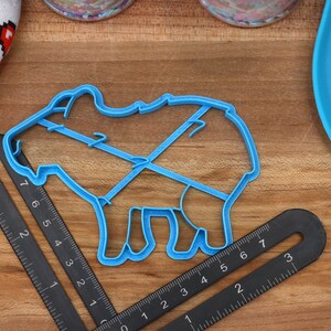 May include: A blue plastic cookie cutter shaped like a pig. The cutter is on a wooden surface with a ruler next to it.