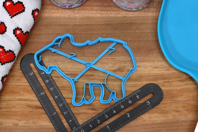 May include: A blue plastic cookie cutter shaped like a pig. The cutter is on a wooden surface with a ruler next to it.