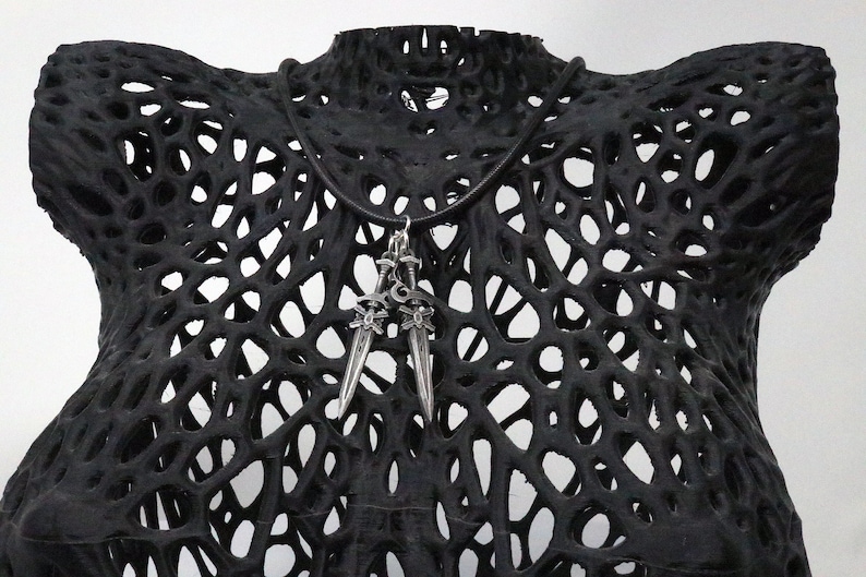 May include: A silver pendant necklace featuring two sword charms. The necklace is displayed on a black, open-weave bust form. The swords have ornate handles and pointed blades. The bust form has a unique, porous design.