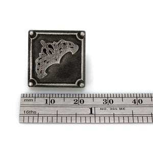 May include: A square, antique silver-toned metal stamp with a decorative, stylized floral design. The stamp is approximately 1 inch square. A metal ruler is in the foreground, providing scale.