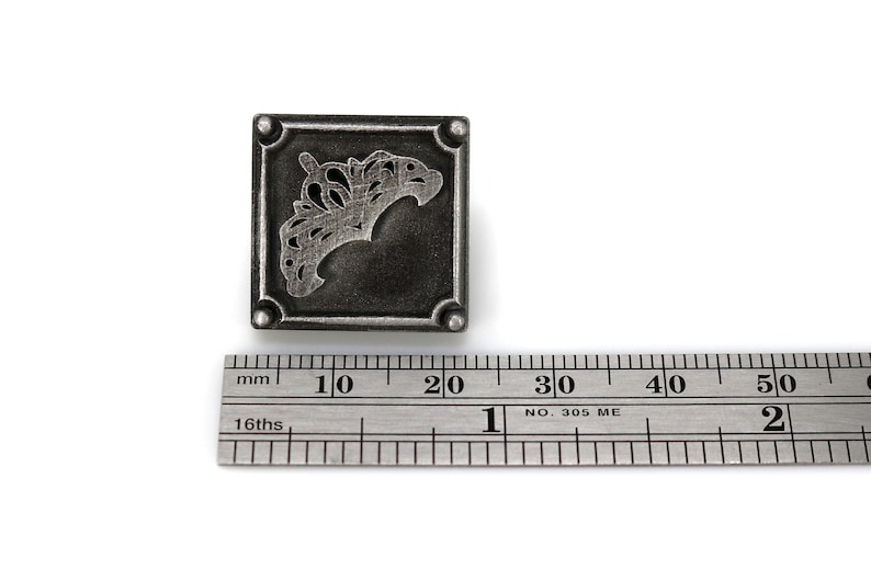 May include: A square, antique silver-toned metal stamp with a decorative, stylized floral design. The stamp is approximately 1 inch square. A metal ruler is in the foreground, providing scale.