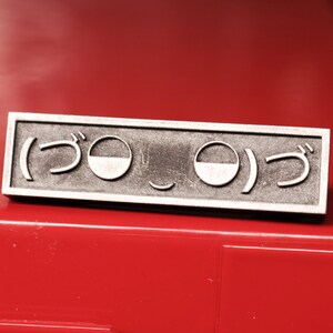 May include: A silver metal pin with a Japanese character and a smiley face design. The pin is on a red surface.