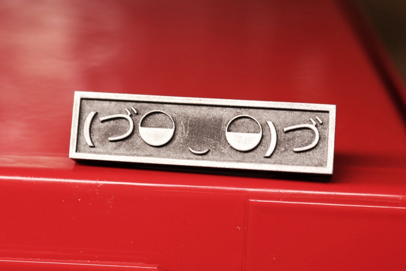 May include: A silver metal pin with a Japanese character and a smiley face design. The pin is on a red surface.