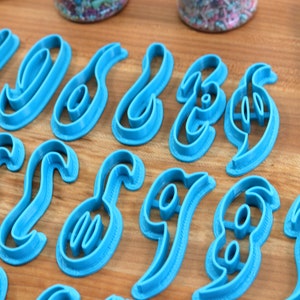 May include: A set of 26 blue plastic cookie cutters shaped like cursive letters of the alphabet. The cutters are arranged in two rows on a wooden surface.