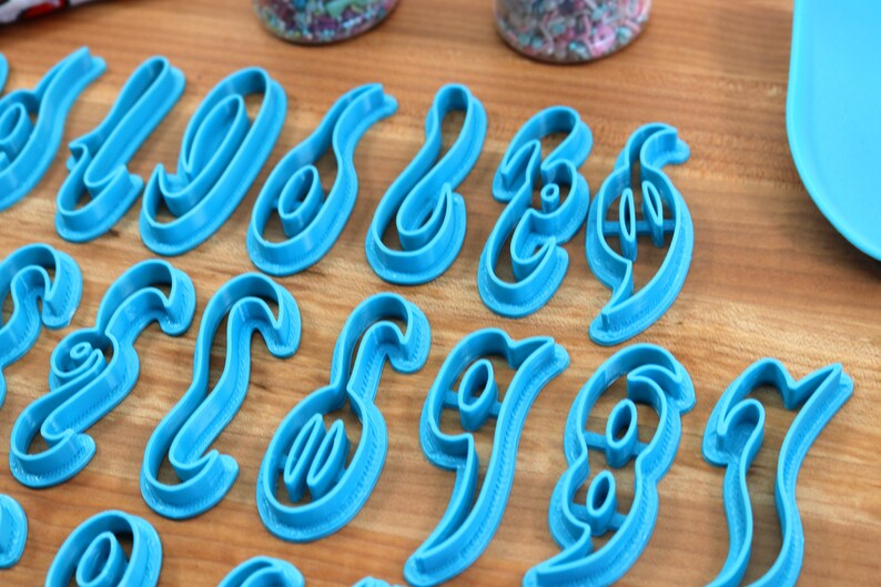 May include: A set of 26 blue plastic cookie cutters shaped like cursive letters of the alphabet. The cutters are arranged in two rows on a wooden surface.