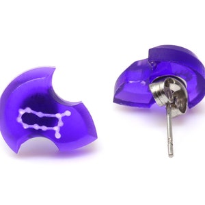 May include: A pair of purple stud earrings with a white Gemini zodiac symbol. The earrings have a silver post and butterfly back.