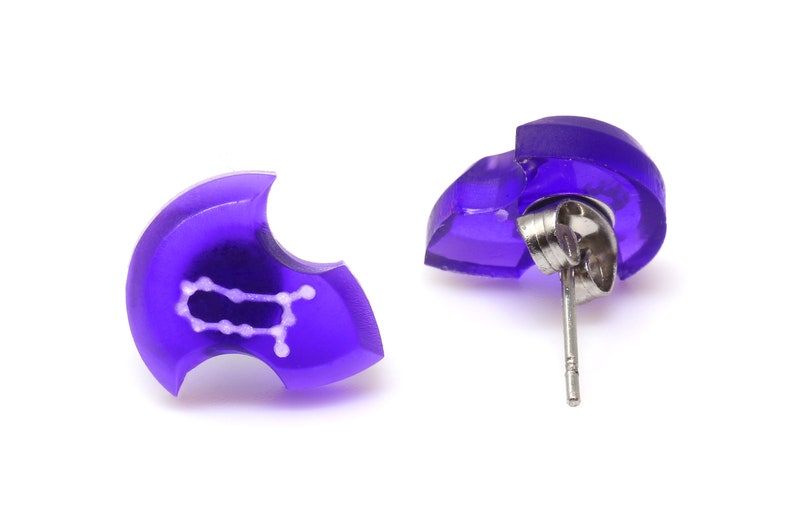 May include: A pair of purple stud earrings with a white Gemini zodiac symbol. The earrings have a silver post and butterfly back.