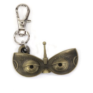 May include: A gold-colored metal keychain featuring a stylized pair of eyes with a small antenna on top. The keychain has a silver metal clasp.