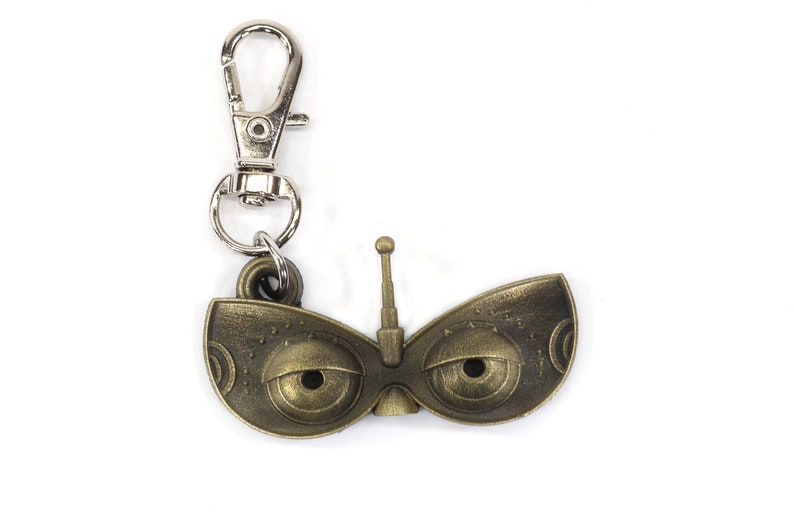May include: A gold-colored metal keychain featuring a stylized pair of eyes with a small antenna on top. The keychain has a silver metal clasp.