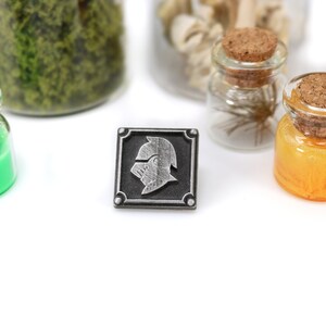 May include: A silver metal pin with a black background and a silver knight's helmet design. The pin is surrounded by a silver border.