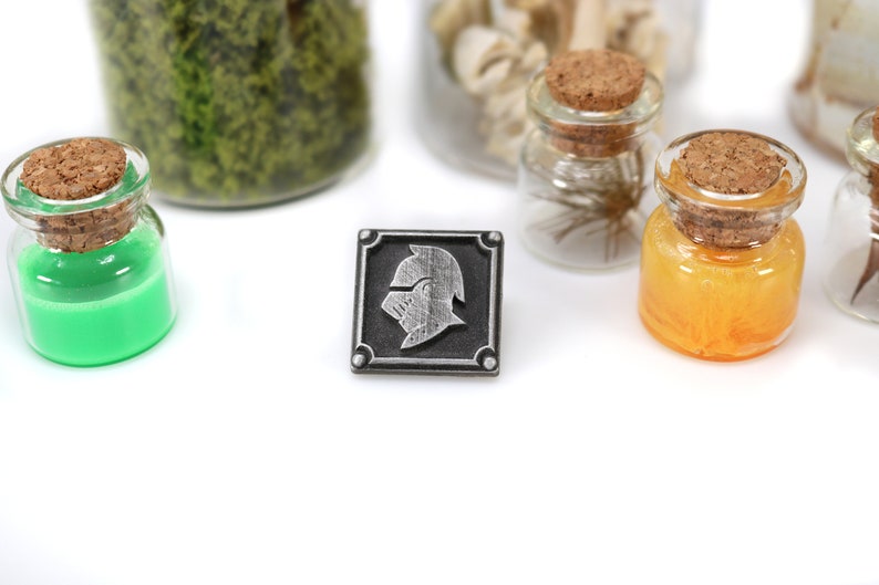 May include: A silver metal pin with a black background and a silver knight's helmet design. The pin is surrounded by a silver border.