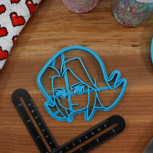 Pu&ograve; includere: Un tagliapasta in plastica blu a forma di personaggio dei cartoni animati con un'espressione seria. Il personaggio ha i capelli corti e spettinati e indossa un abbigliamento semplice.