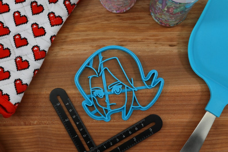 Pu&ograve; includere: Un tagliapasta in plastica blu a forma di personaggio dei cartoni animati con un'espressione seria. Il personaggio ha i capelli corti e spettinati e indossa un abbigliamento semplice.
