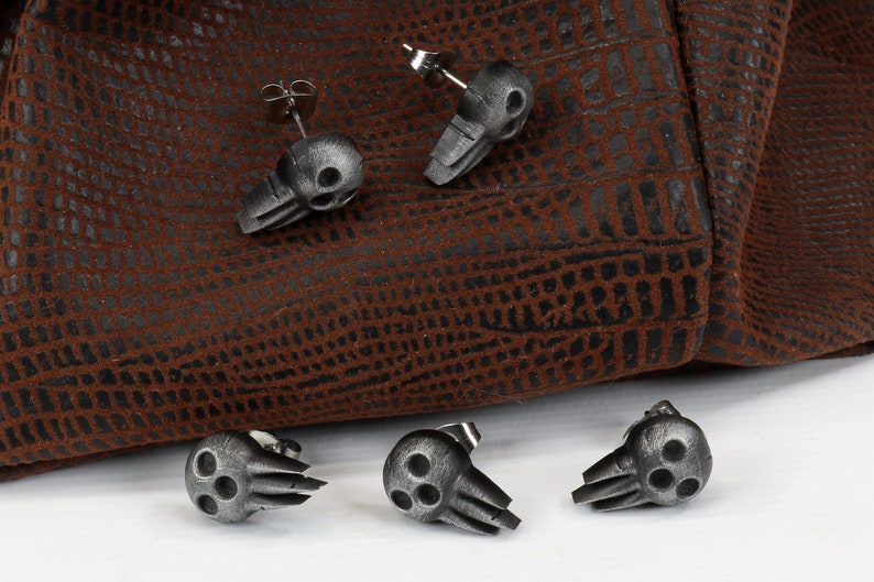 Peut inclure: Cinq boucles d'oreilles en forme de cr&acirc;ne argent&eacute;es avec trois orbites sur un fond en cuir marron.