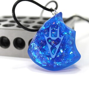 May include: A blue resin pendant necklace with a white hand-shaped design and iridescent glitter embedded in the resin. The pendant is shaped like a jagged rock.