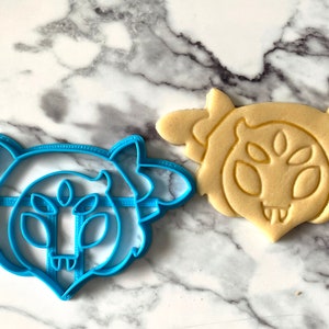 May include: A blue plastic cookie cutter shaped like a cartoon monster with three eyes and a toothy grin. Next to it is a cookie baked in the shape of the monster.