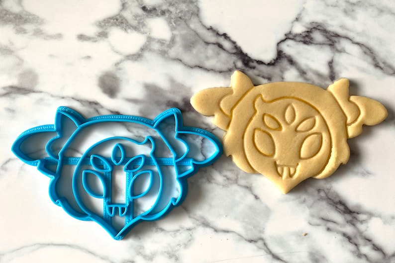 May include: A blue plastic cookie cutter shaped like a cartoon monster with three eyes and a toothy grin. Next to it is a cookie baked in the shape of the monster.