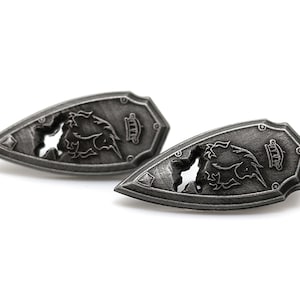 May include: Two silver-toned metal shield-shaped earrings with a detailed design of a dragon with its wings spread out. The dragon is partially cut out of the metal, creating a negative space effect.