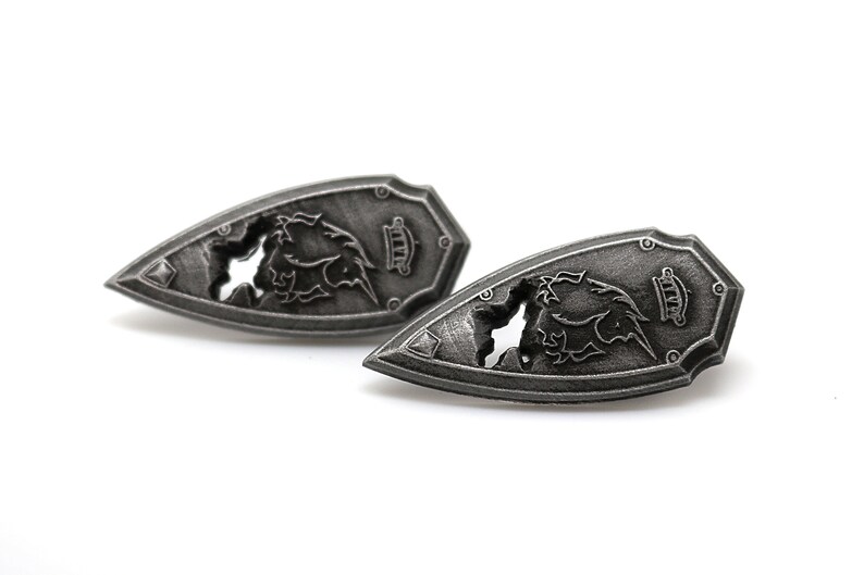 May include: Two silver-toned metal shield-shaped earrings with a detailed design of a dragon with its wings spread out. The dragon is partially cut out of the metal, creating a negative space effect.