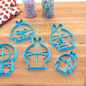 May include: Five blue plastic cookie cutters shaped like cartoon bunnies with different facial expressions. The cutters are on a wooden surface.