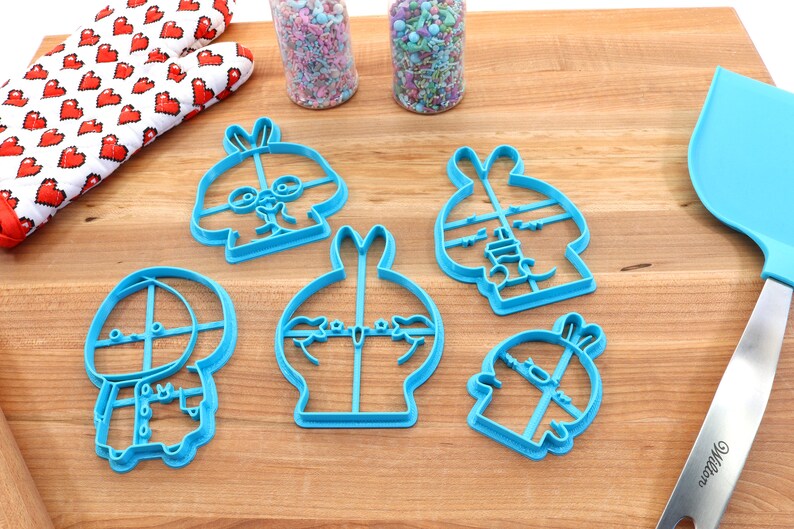May include: Five blue plastic cookie cutters shaped like cartoon bunnies with different facial expressions. The cutters are on a wooden surface.