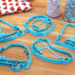 May include: Five blue plastic cookie cutters shaped like a sailboat, a car, a banana, a swing set, and a donut. The cutters are on a wooden surface.