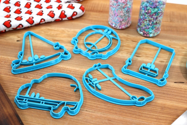 May include: Five blue plastic cookie cutters shaped like a sailboat, a car, a banana, a swing set, and a donut. The cutters are on a wooden surface.
