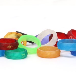 May include: A collection of colorful, geometric rings with different symbols and designs. The rings are made of a translucent material and are stacked on a white surface.