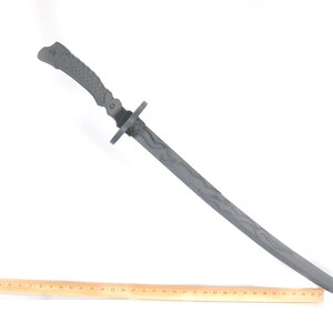 Może przedstawiać: Szary miecz katana z teksturowanym uchwytem i wzorem ostrza. Miecz jest ustawiony pod kątem w poprzek ramy, z drewnianą linijką umieszczoną poniżej dla skali. Ostrze miecza ma wz&oacute;r falisty.