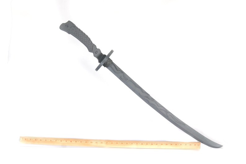 Może przedstawiać: Szary miecz katana z teksturowanym uchwytem i wzorem ostrza. Miecz jest ustawiony pod kątem w poprzek ramy, z drewnianą linijką umieszczoną poniżej dla skali. Ostrze miecza ma wz&oacute;r falisty.