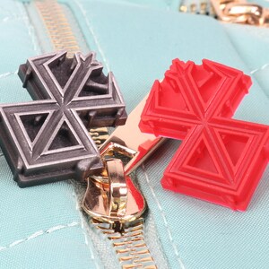 May include: Two zipper pulls, one silver and one red, in the shape of a star with a geometric design. The zipper pulls are attached to a light blue zipper.