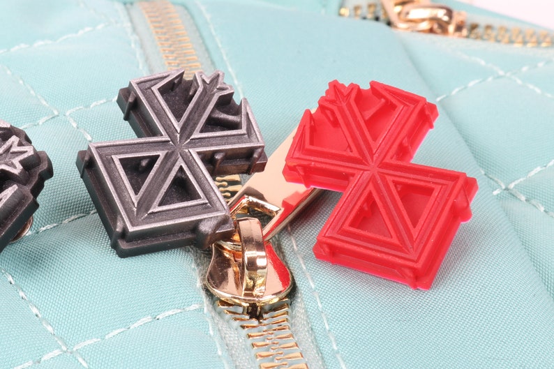 May include: Two zipper pulls, one silver and one red, in the shape of a star with a geometric design. The zipper pulls are attached to a light blue zipper.