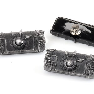May include: Three silver-coloured metal pins with intricate designs. The pins are shaped like a rectangular box with a circular detail in the centre. The pins have a black backing with a pin back.