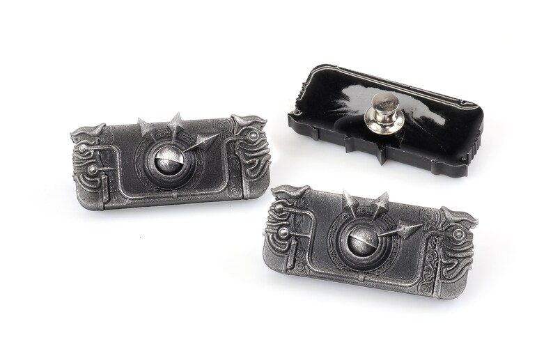 May include: Three silver-coloured metal pins with intricate designs. The pins are shaped like a rectangular box with a circular detail in the centre. The pins have a black backing with a pin back.
