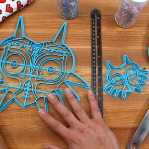 May include: Two blue plastic cookie cutters shaped like the iconic Legend of Zelda mask, one large and one small, on a wooden surface.