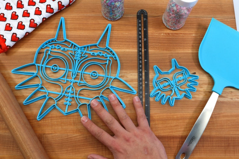 May include: Two blue plastic cookie cutters shaped like the iconic Legend of Zelda mask, one large and one small, on a wooden surface.