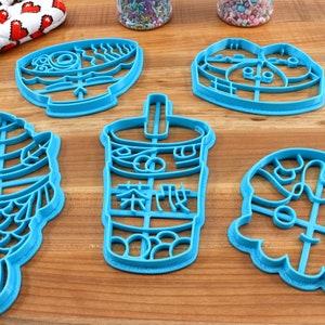 May include: A collection of six turquoise plastic cookie cutters in various shapes, including a fish, a bubble tea cup with Chinese characters, and other food-related designs. The cutters are arranged on a wooden surface.