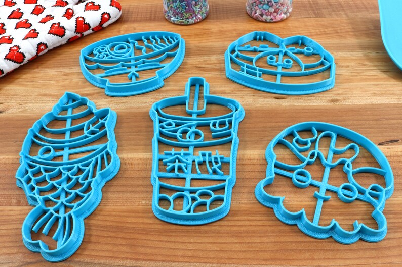 May include: A collection of six turquoise plastic cookie cutters in various shapes, including a fish, a bubble tea cup with Chinese characters, and other food-related designs. The cutters are arranged on a wooden surface.