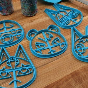 May include: Five blue plastic cookie cutters shaped like animal faces. The cutters are on a wooden surface. The animal faces include a bear, a cat, and three different variations of a fox.