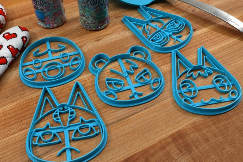 May include: Five blue plastic cookie cutters shaped like animal faces. The cutters are on a wooden surface. The animal faces include a bear, a cat, and three different variations of a fox.