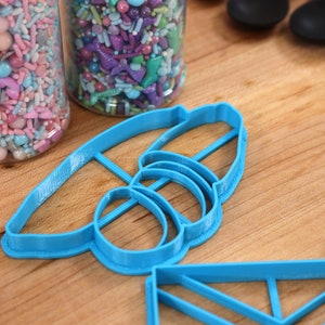 May include: A set of blue plastic cookie cutters shaped like a bee, a rectangle, and a square. The cutters are on a wooden surface.