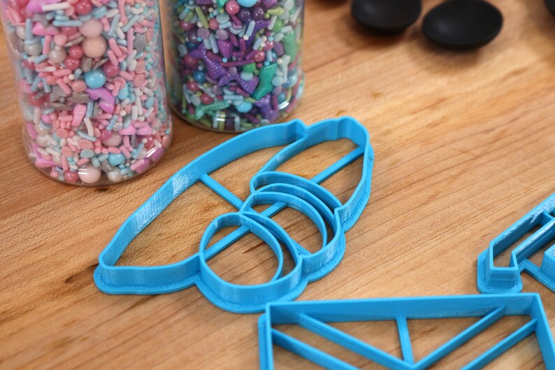 May include: A set of blue plastic cookie cutters shaped like a bee, a rectangle, and a square. The cutters are on a wooden surface.