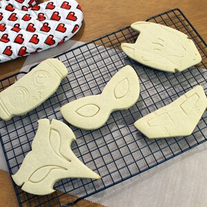 May include: An assortment of unbaked cookies in various shapes, including a bottle, mask, and geometric designs, are arranged on a wire cooling rack. A red and white oven mitt with a pixelated heart pattern is in the background.