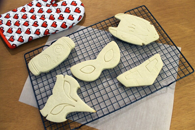 May include: An assortment of unbaked cookies in various shapes, including a bottle, mask, and geometric designs, are arranged on a wire cooling rack. A red and white oven mitt with a pixelated heart pattern is in the background.