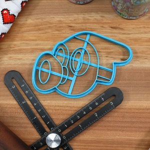 May include: A blue plastic cookie cutter shaped like a cartoon character with a large nose and eyes. The cutter is on a wooden surface with a ruler and rolling pin.
