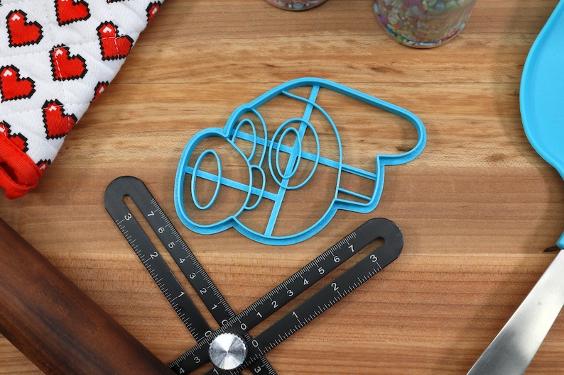 May include: A blue plastic cookie cutter shaped like a cartoon character with a large nose and eyes. The cutter is on a wooden surface with a ruler and rolling pin.