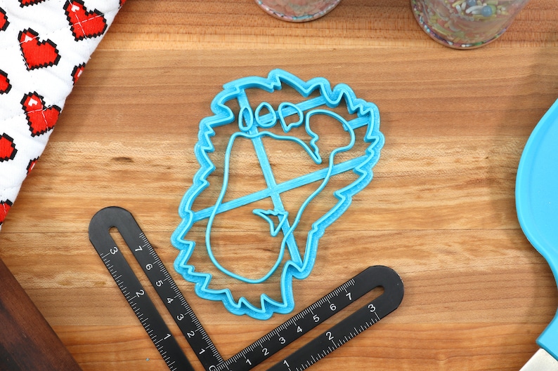 May include: A blue plastic cookie cutter shaped like a footprint with a jagged edge. The cutter is on a wooden surface.