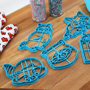 Puede incluir: Cinco cortadores de galletas de pl&aacute;stico azul con forma de mapaches y un cubo de palomitas de ma&iacute;z. Los cortadores est&aacute;n sobre una superficie de madera.