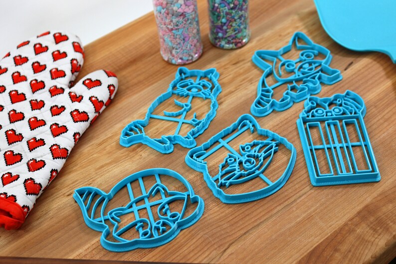 Puede incluir: Cinco cortadores de galletas de pl&aacute;stico azul con forma de mapaches y un cubo de palomitas de ma&iacute;z. Los cortadores est&aacute;n sobre una superficie de madera.